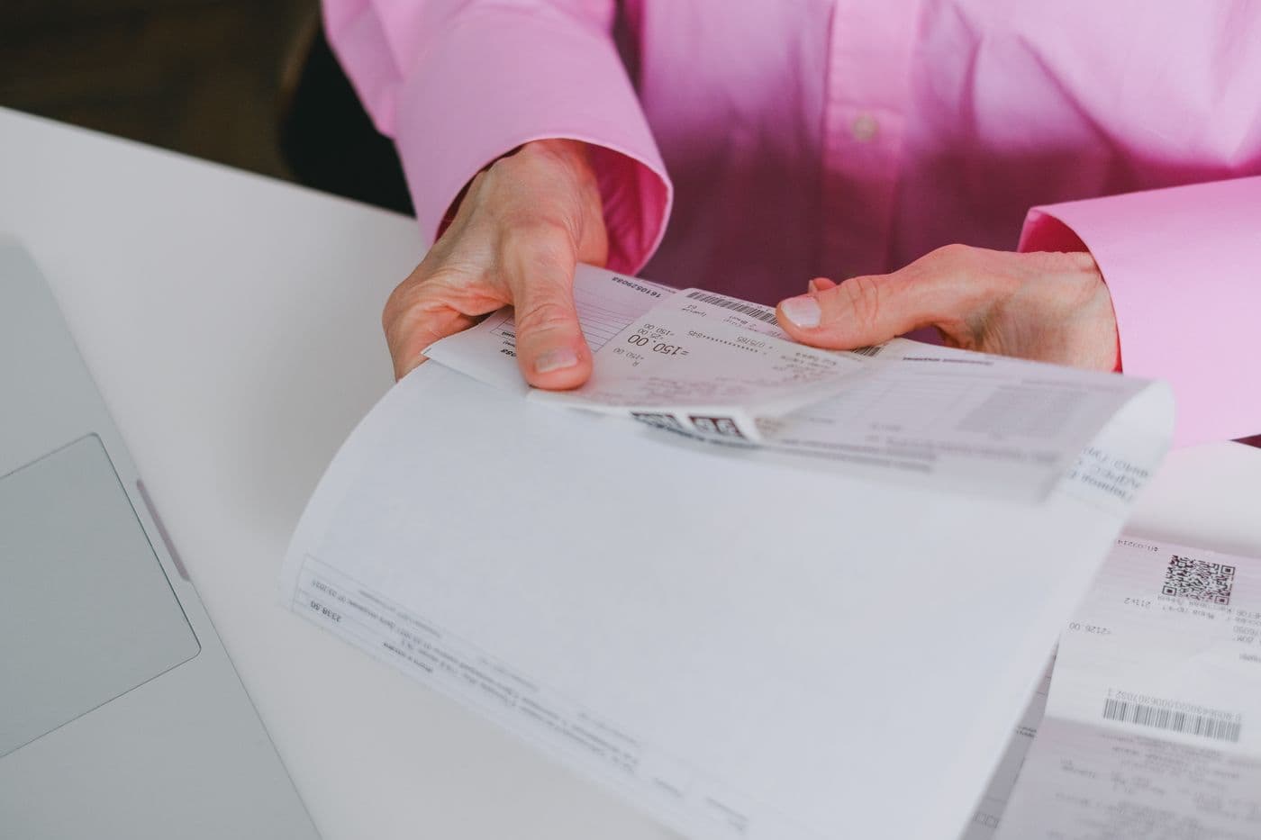 Mains classant des factures et des justificatifs de dépenses sur un bureau blanc, à côté d'un ordinateur portable