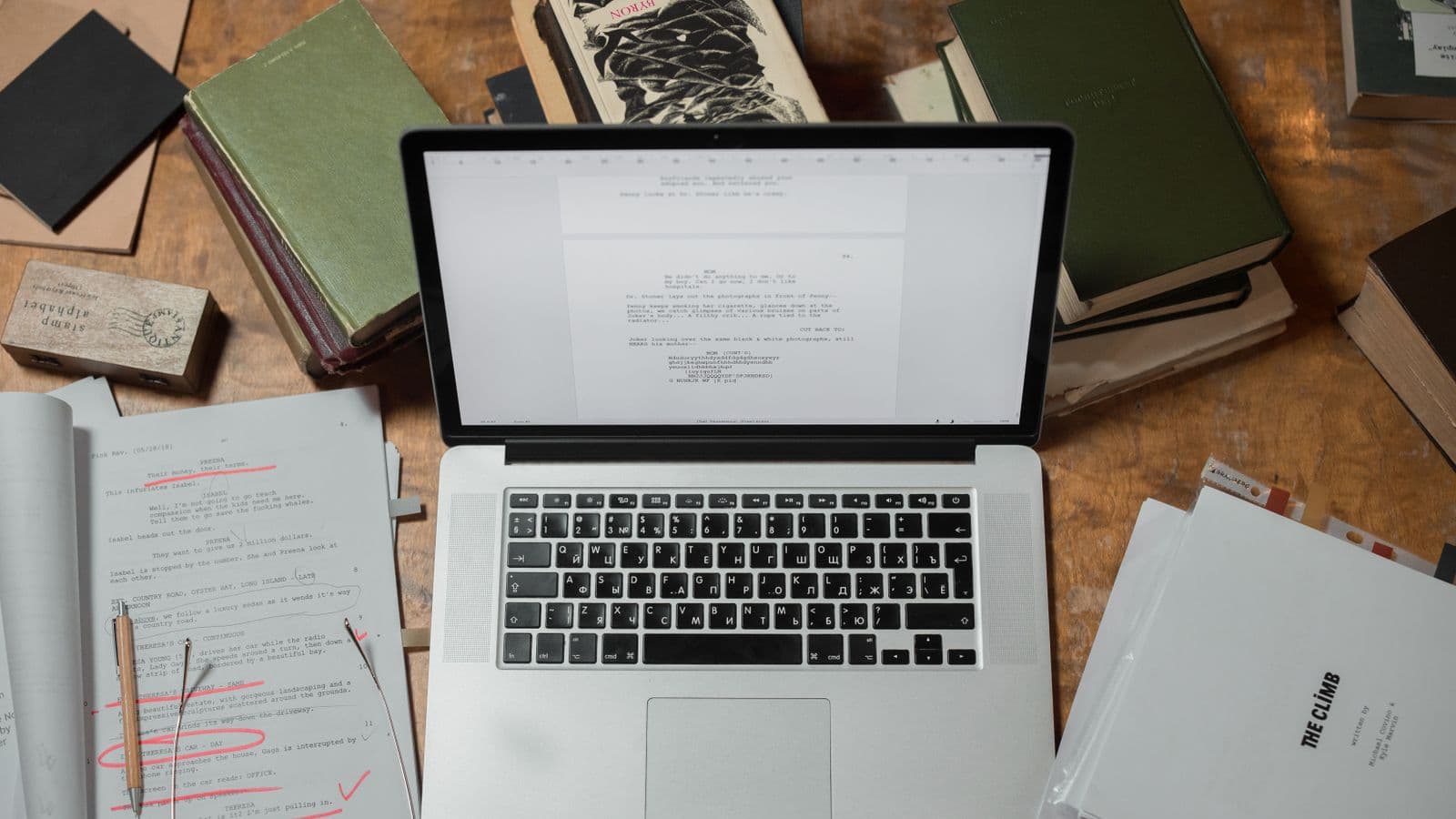 A screenplay marked up in red pen beside a laptop showing script pages and a stack of books on a wooden desk