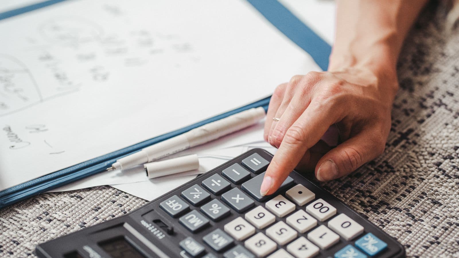 Une main pose un total sur une calculatrice, à côté de notes manuscrites sur un porte-bloc
