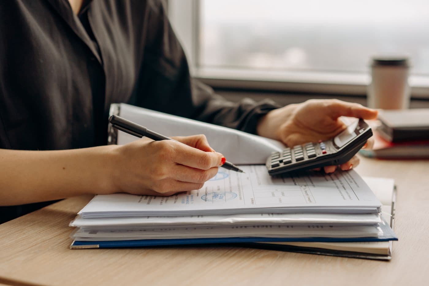 Hands holding a calculator over a stack of paperwork and files on a wooden desk