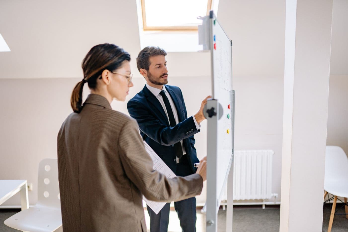 Deux personnes discutent devant un tableau blanc dans un bureau lumineux, une liste imprimée à la main