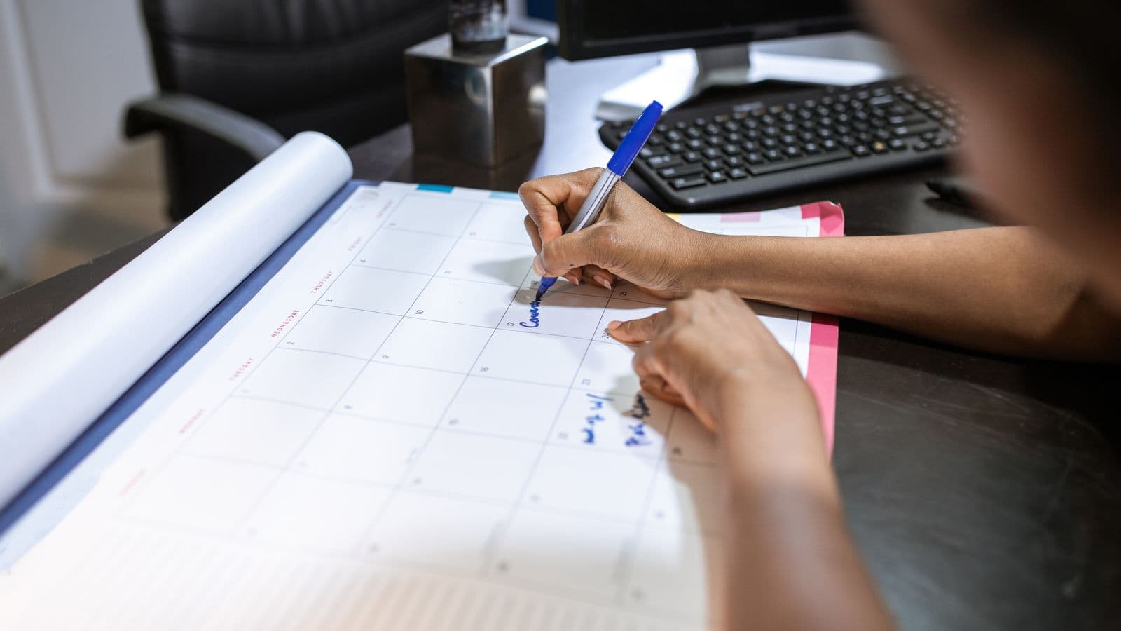 Mains annotant au stylo un planning imprimé posé sur un bureau, à côté d'un clavier d'ordinateur
